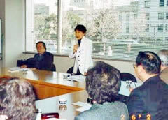長方形に並べられた机に着席する要請参加者と、席で立って話している川田悦子衆議院議員。