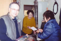 玄関先で、訪問者と訪問先の方の3人。お見舞い金と編み物を手渡している場面