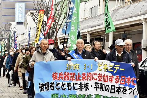 「核兵器禁止条約に参加する日本に」など書かれた横断幕を持ち、道路を行進するビキニデー集会参加者たち。後ろには参加した団体ののぼりや旗もみえており、人の列が続いている。