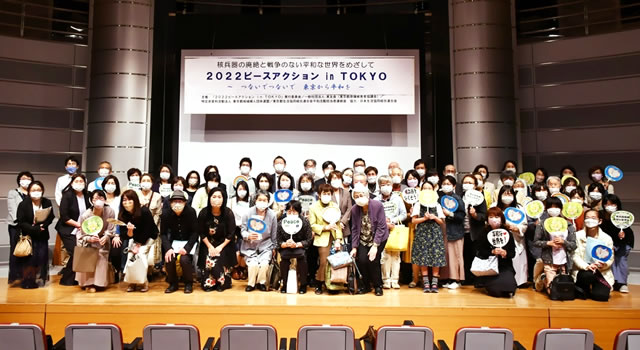 舞台の上に密集して並ぶ参加者たち。全員がマスクを着用しており、なにかメッセージが描かれたうちわを持っている人達が多い。天井からは「2022 ピースアクション in TOKYO」など大きく横書きれた看板が天井から提げられている。