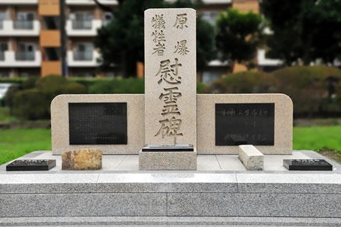 慰霊碑を正面から撮った写真。中央に「原爆犠牲者 慰霊碑」と縦書きの文字が彫られた高い石の両袖に、碑文などが刻まれた石がはめ込まれている。