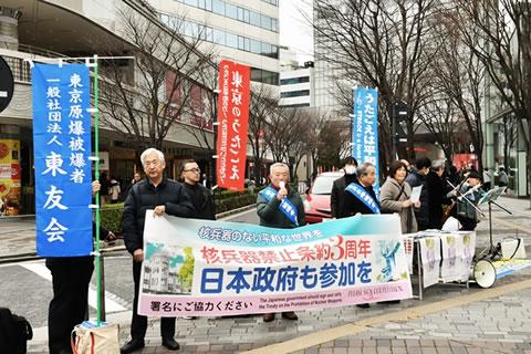 「核兵器禁止条約発効3周年 日本政府も参加を」と書かれた日本原水協の横断幕を持って街頭に立つ参加者たち。横断幕には広島の原爆ドーム、長崎の平和祈念像の写真もプリントされている。「東京原爆被爆者 一般社団法人 東友会」ののぼり、「東京のうたごえ」と書かれたのぼりを立てている参加者もいる。被爆者はたすきをかけている。参加者の一人がマイクを持ち、拡声器を使って通る人たちに署名を呼び掛けている。