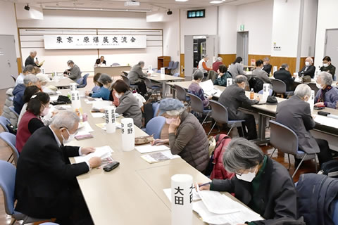 前方に「東京・原爆展交流会」と大きく横書きされた看板の下げられた広い部屋。並べられた着席した参加者が、手元の資料を見るなどしながら、演台で話す人の話を聴いている。