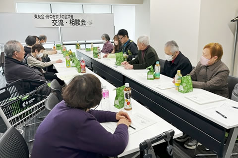 向き会う形で2列並べられた長机に着席した参加者たち。前方のホワイトボードに、「東友会・府中きすげの会共催 交流・相談会」と大きく書かれた紙が張られている。