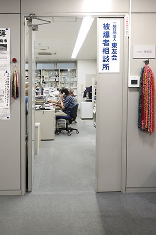 廊下から写した、東友会入り口。「東友会 被爆者相談所」の掲示がある。ドアは室内に向いて開いており、左右の壁には千羽鶴などがさげられている。