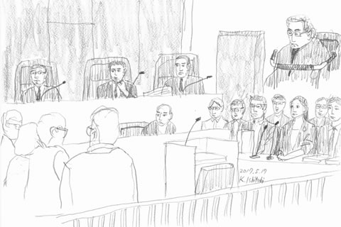 法廷のスケッチ。被爆者側の席、着席している弁護士の前にマイクが置かれている。国側の出席者と裁判官も描かれている。被爆者側の席の上余白に、着席した原告山本さんが描かれている。