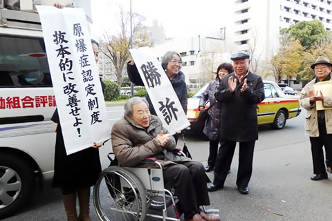 車いすに乗った笑顔の山本さんと、両脇で「勝訴」「原爆症認定制度 抜本的に改善せよ」と書かれた紙を掲げる人、周りに拍手する支援者