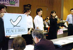 模造紙くらいの大きさのポスターを二人がもち、他のメンバーとともに並んで立っている。一人がマイクを持って話をしている。シンボルマークは、「翼を広げた鳩」と「ハートの形」を合わせて図案化したもの。輪郭は一本の線のみで描かれている。