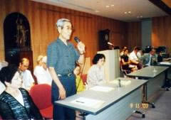 長机の席で立ってマイクを使い話す要石謙次さん。横に並べられた長机やその後ろにも参加者が座っている。