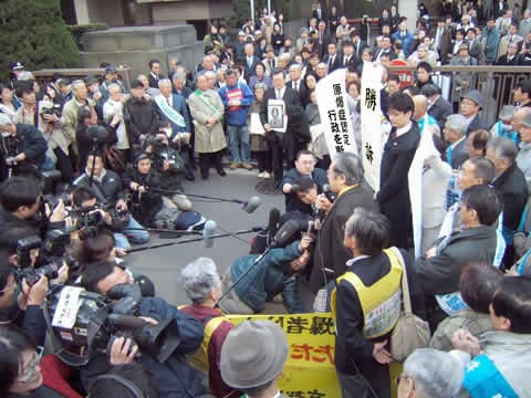 東京地裁前を埋める原告・弁護団・支援者とマスコミの取材陣。多数のカメラとマイクが原告に向けられている。弁護士は「勝訴」の文字などを掲げている。
