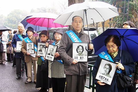厚生労働省前で、雨の中傘を差し、亡くなった集団訴訟原告の遺影を掲げて訴える被爆者たち。