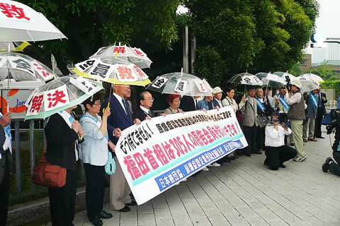 総理官邸前で、「全員救済」などの文字を貼ったビニール傘を差し、横断幕を掲げて訴える人たち。