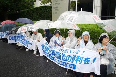 厚生労働省前でカッパを着、「全面解決」など貼り付けたビニール傘を差し、横断幕を広げて座り込みに参加する原告・被爆者たち