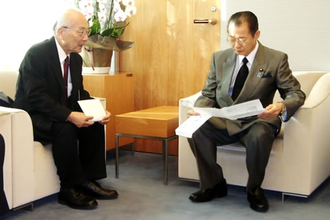 官房長官に向かって話をしている日本被団協の田中煕巳事務局長と、紙を広げて目を落としながら聞く河村建夫官房長官。2名とも椅子に座っている。