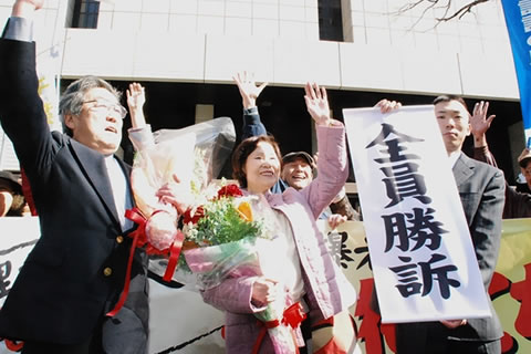 「全員勝訴」の文字を掲げる弁護士と、その隣に並ぶ原告ら。手渡された花束を抱え片腕を上にあげた原告の後ろで、支援者たちも手を挙げて喜びを表している。