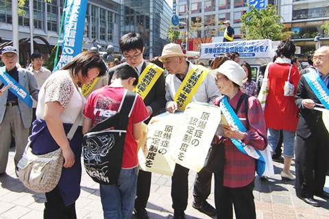 渋谷駅ハチ公前広場で、たすきを掛けて署名判を持つ被爆者や支援者たち。署名に応じる家族連れも。