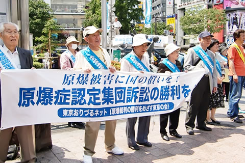 5月、渋谷ハチ公前ひろばで、東友会のたすきを掛け、「原爆症認定集団訴訟の勝利を」と書かれた横断幕を掲げて立つ被爆者たち。