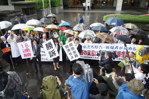 東京高裁前で、雨の中傘を差しながら横断幕などを持つ支援者たち。その前で弁護士が「勝訴」「18連勝」など書かれた紙を掲げている。その手前に、カメラやマイクを構えたマスコミの取材陣。