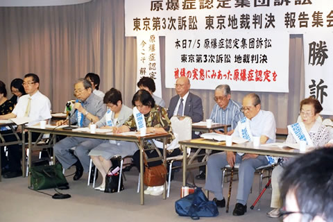 並べられた机に着席する原告や弁護士たち。その後ろの壁に、「東京地裁判決報告集会」などの横幕、「勝訴」の文字などが掲げられている。