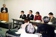 会場前方、演壇に立つ田川会長代行。その横に長机が並べられており、都側の出席者が座っている。
