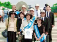 広島の平和記念公園で、原爆ドームを遠景に集まって立つ代表団。何人かはたすきを掛け、一人は故・田川会長の遺影を持っている。