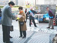 新宿駅前の歩道でチラシを配り、歩行者と話をする参加者たち。歩道などが貼られた大きい紙も広げられている。