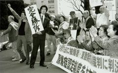 「完全勝訴」の文字を掲げる弁護士と、その周辺で拍手をしたりこぶしを挙げる被爆者、支援者ら。被爆者と支援者は、横断幕を前にして座っている人と、立っている人がいる。2004年3月31日、東京地方裁判所前で、共同通信社撮影
