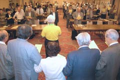 会場前方の壇上で立っている人たちの背後から撮られた写真。写真奥では並べられた机の席で参加者が立っている。