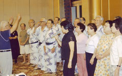 ホールで立って並び、指揮をする人に合わせて歌う参加者ら。浴衣を着ている人も。