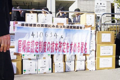 2月18日、厚生労働省前に積み上げられた、署名の入った段ボール箱。側面に、「原爆症認定制度の抜本改定を求める署名」と書かれた横断幕がかけられている。