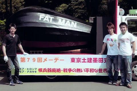 「ファットマン」実物大模型を荷台に乗せたトラックと、3人の若者たち。荷台には「第79回メーデー 東京土建隅田支部」など書かれた横断幕が貼られている。模型は、実物のような楕円球型ではなくナス型で、尾翼も実物よりずっと小さい。
