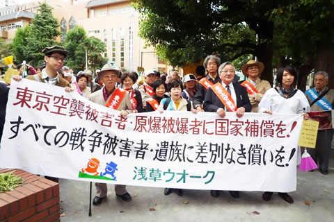 「ウォーク」の先頭に掲げられた「東京空襲被害者・原爆被爆者に国家補償を！全ての戦争被害者・遺族に差別なき償いを！」の横断幕。横断幕を持つ人たちの後ろに人の列が続いている。