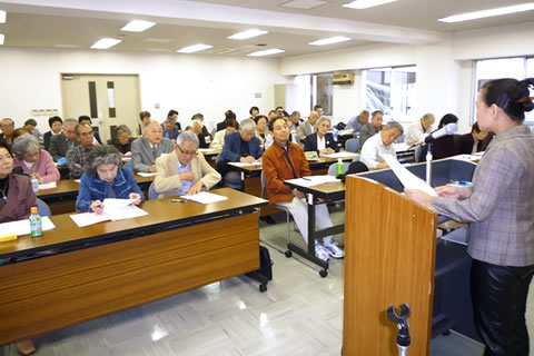 並べられた机に着席し講演を聞く参列者たちと、講師の園田久子医師。
