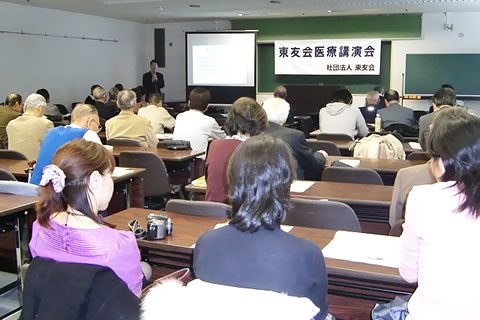 前方に投影用のスクリーンがおろされた会場、並べられた机に着席し講演を聞く参加者たち。