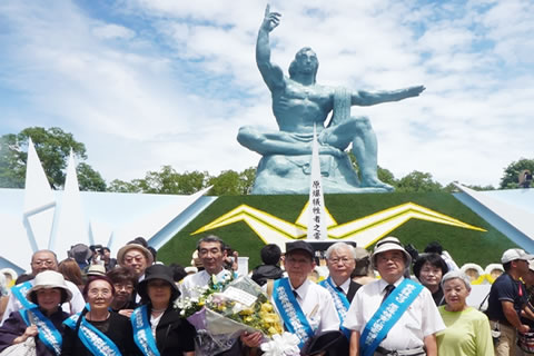 たすきを掛けた東友会代表団の集合写真。花束を持つ方もいる。背景に平和祈念像。