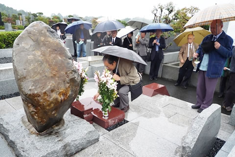 傘をさして墓の前に並ぶ参加者たち。1人はしゃがんで墓に花を供えている。