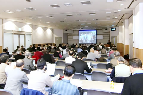 並べられた机に着席し講演を聞く参加者たち。会場前方のスクリーンにスライドが投影されている。