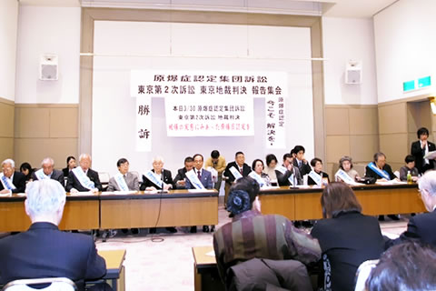 原告団・弁護団が会場前方の机に着席している。集会参加者たちが写真手前に写っている。