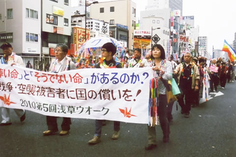 「尊い命とそうでない命があるのか？」「空襲被害者に国の償いを」と書かれた横断幕を先頭に掲げて進む行進。千羽鶴、文字の書かれた傘、旗などを持っている人もいる。