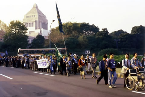 国会議事堂を背景にすすむ国会請願行進。長く続く人の列が車道を進んでいる。横断幕や旗などを持った人もいる。