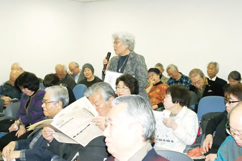 席で立って発言する参加者と、周囲の席に座る参加者たち。