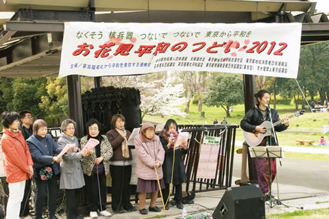 第五福竜丸のエンジンの前に立ち、ギター演奏にあわせ歌う人たち。頭上には「お花見平和のつどい2012」と書かれた横幕が提げられている。