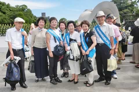 広島平和記念公園にて、たすきをかけた代表団の集合写真。背景とおくに原爆ドーム。