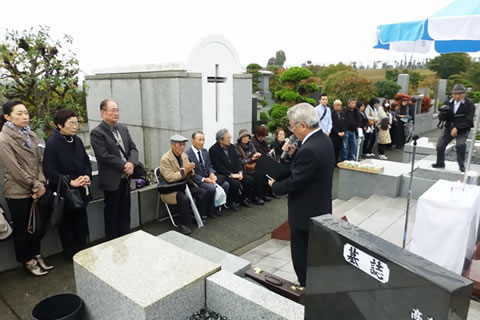 「原爆被害者の墓」の周りに集まった参加者は、椅子に腰かけたり立ったりしながら話を聞いている。