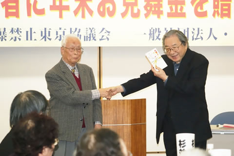 壇上で握手する都丸哲也さんと山本英典さん。山本さんがもう一方の手で見舞金を持っている。