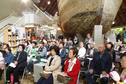 展示された第五福竜丸船体の下、舳先側に並べられた椅子に座る参加者たち。