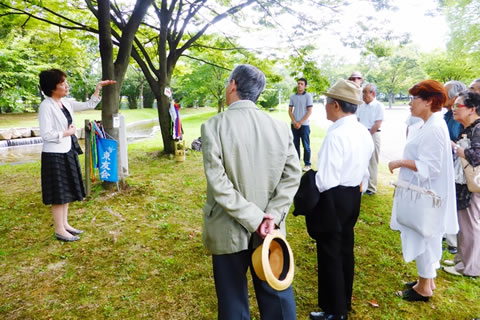 広島市中央公園内、2本のケヤキの前に立つ東友か代表たち。並んだ人たちに向き合って立つ一人が、身振り手振りを交えて話をしている。