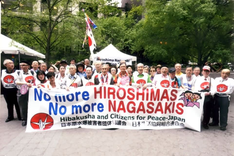 公園の一角のような場所で、「No More HIROSHIMAS No more NAGASAKIS」と書かれた横断幕を広げて立つ、40人ほどの代表団の集合写真。背後にはテントがあり、その奥に広葉樹の林が見える。