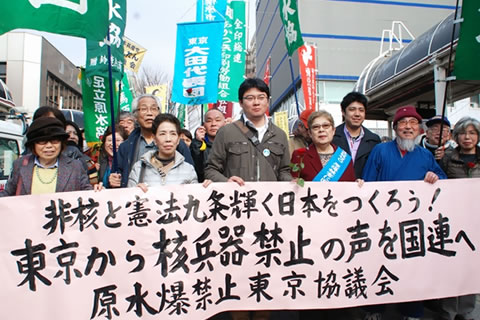 「非核と憲法9条輝く日本をつくろう！東京から核兵器禁止の声を国連へ」と書かれた横断幕を7人が横に並んで掲げ、後ろにつながる行進参加者は団体の旗などを掲げている。笑顔の墓参行進参加者たち。
