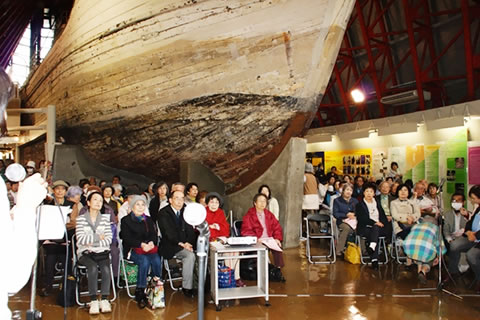 第五福竜丸展示館内、船体を囲むように設置された座席に座り、報告に耳を傾ける人たち